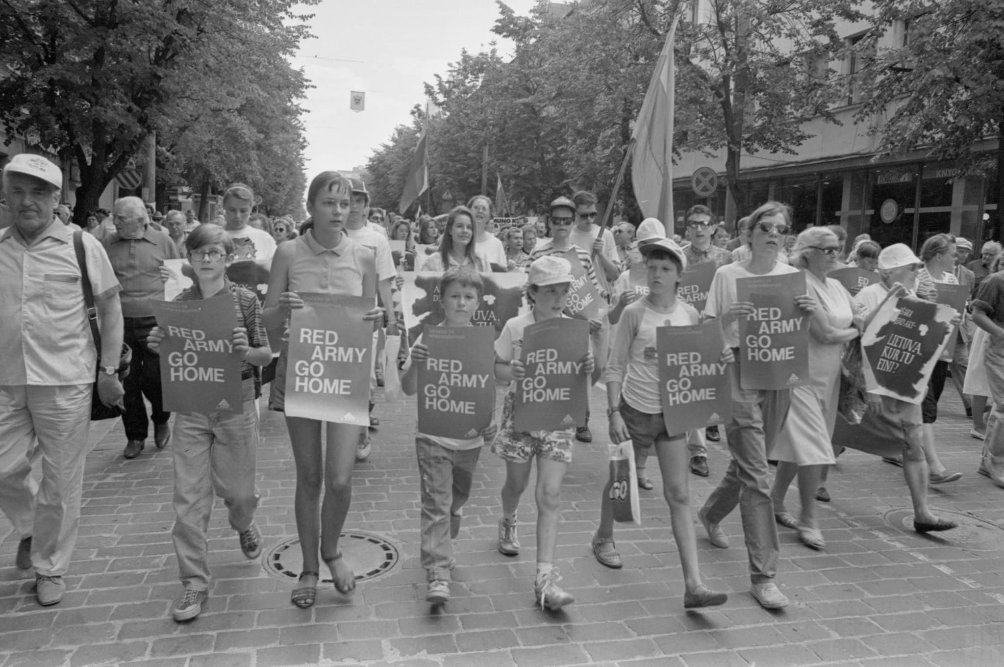 A procession of people in Vilnius main street. 6 kids in front of the procession carry red posters with 'Red army go home' in white.